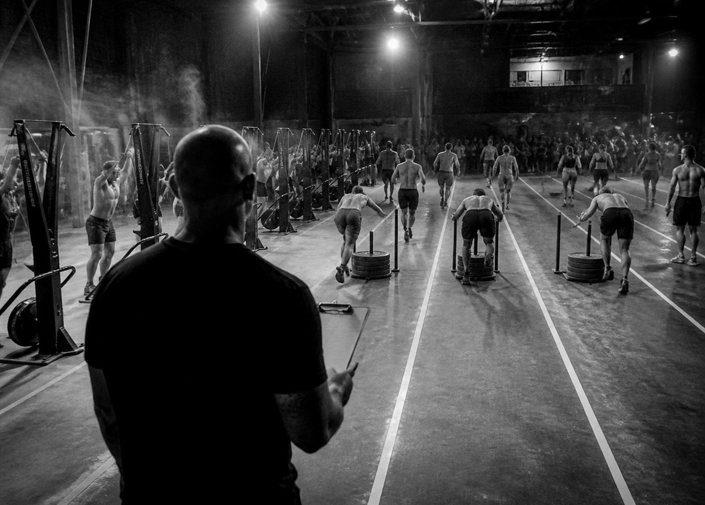 A gym owner with a clipboard watching athletes run a lane-by-lane fitness simulator event
